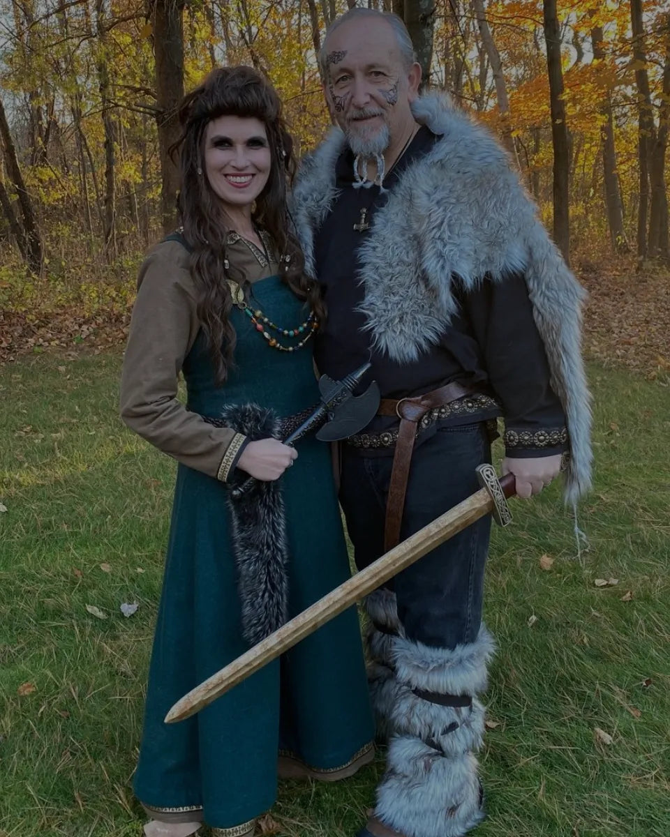 A couple dressed in authentic Viking-inspired clothing, including a green tunic and fur-lined mantle, posing outdoors with custom-crafted artisan axes and swords.