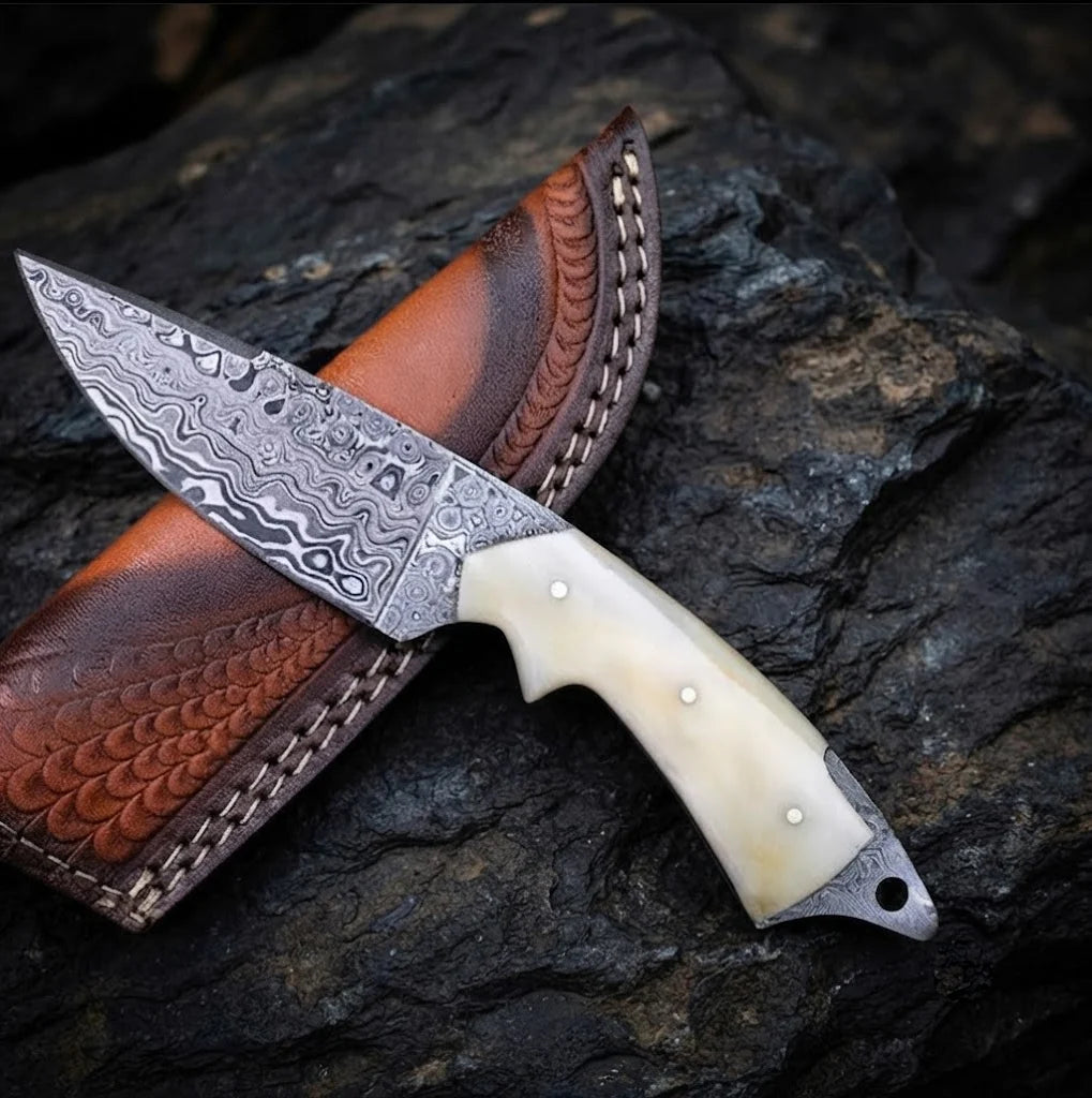 A premium handmade Damascus steel skinner knife featuring a smooth camel bone handle, 512-layer steel construction, and a custom-fitted leather sheath on a dark stone background.