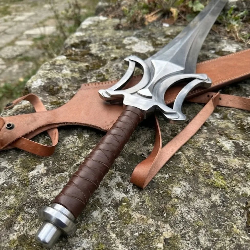 A close-up of a custom hand-forged fantasy short sword featuring a unique steel guard and a brown leather-wrapped handle, resting on a rocky surface with its leather sheath.