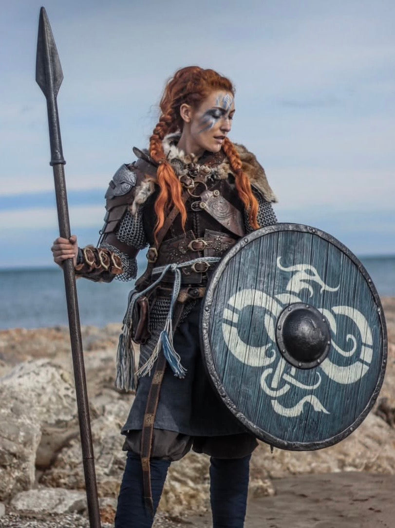 Full-body portrait of a Viking shieldmaiden standing on a rocky beach, wearing detailed leather armor and chainmail, while holding a custom spear and signature dragon-motif shield.
