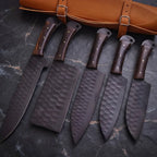 A 5-piece professional kitchen knife set featuring hammered carbon steel blades and dark hardwood handles, displayed with a premium leather storage roll on a dark stone background.