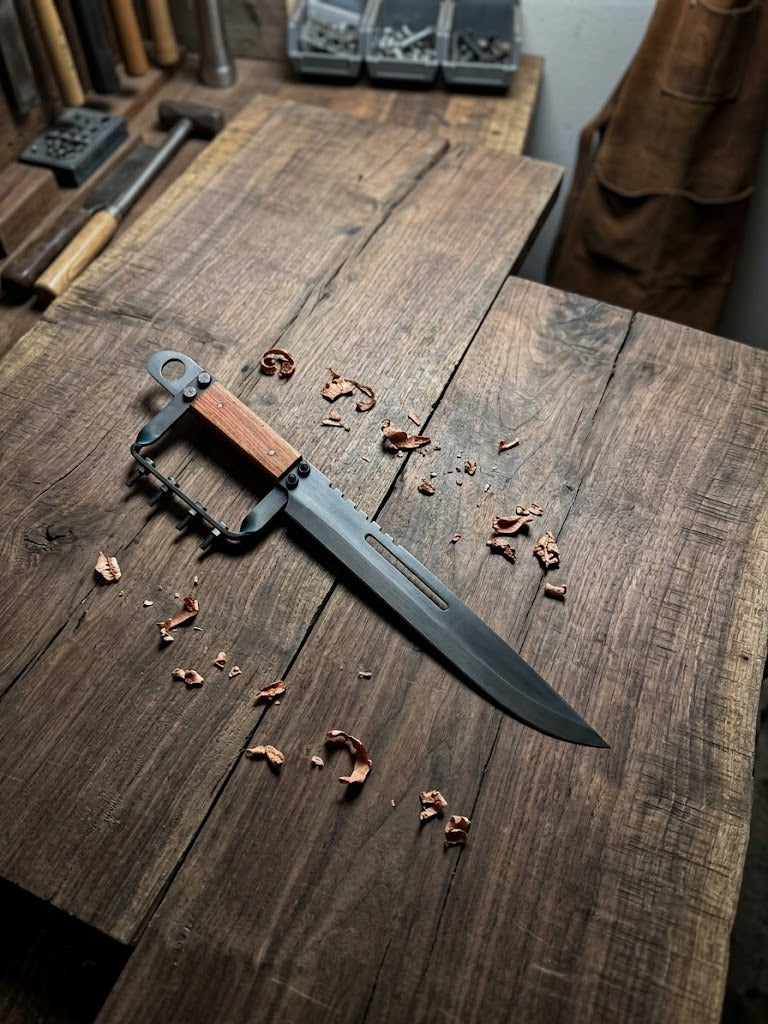 Hand-forged artisan tactical knife featuring a unique guard design and ergonomic wooden grip, set in a professional forge environment.