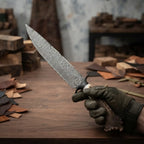 A close-up view of a premium hand-forged Damascus steel knife with a unique antler handle, being held by a gloved hand in a workshop.