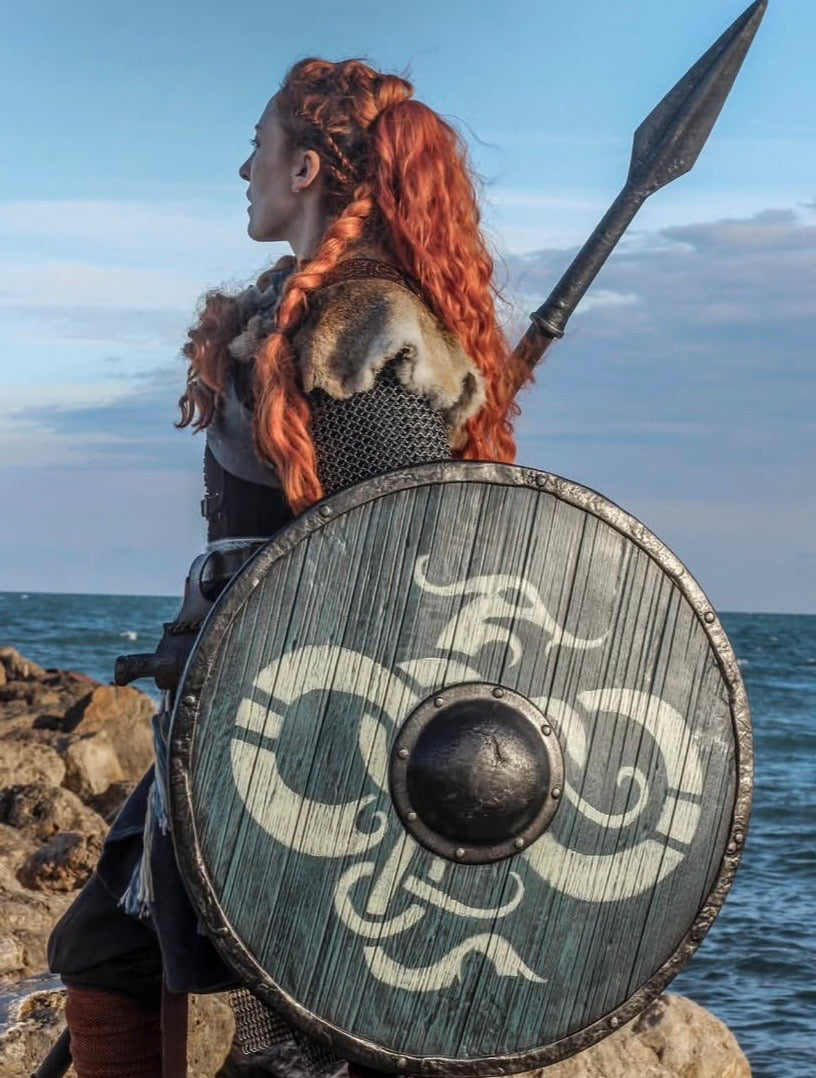 Viking shieldmaiden in profile, equipped with a custom handcrafted dragon-motif shield and battle spear on a rocky coastal landscape.