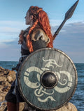 Viking shieldmaiden in profile, equipped with a custom handcrafted dragon-motif shield and battle spear on a rocky coastal landscape.