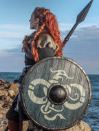 Viking shieldmaiden in profile, equipped with a custom handcrafted dragon-motif shield and battle spear on a rocky coastal landscape.