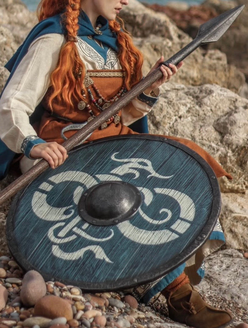 Detailed close-up of a Viking warrior in traditional attire, holding a handcrafted wooden spear and a signature dragon-motif shield while seated on a rocky shore.