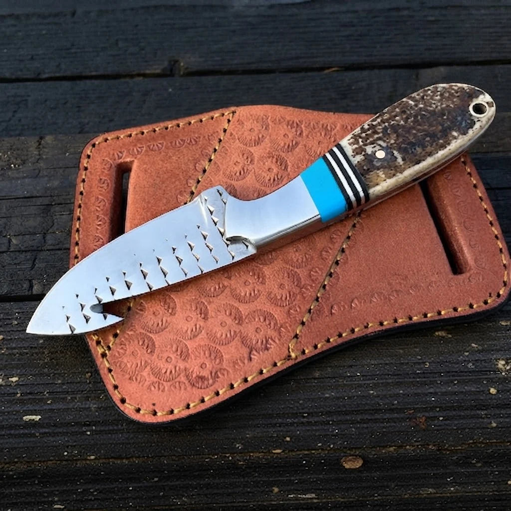 A professional top-down view of a handmade D2 steel cowboy gut hook knife with an intricate file-worked spine, stag antler handle, and turquoise spacers resting on a pancake leather sheath.