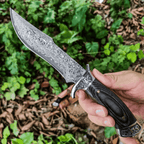 Close-up of a person holding a handcrafted Damascus steel Bowie knife with a highly detailed twist pattern blade, featuring an engraved silver guard and dark wooden handle against a natural outdoor background.