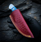 A handmade Damascus steel skinner knife tucked into a beautifully hand-stitched maroon leather sheath, held by a gloved hand against a dark stone background.