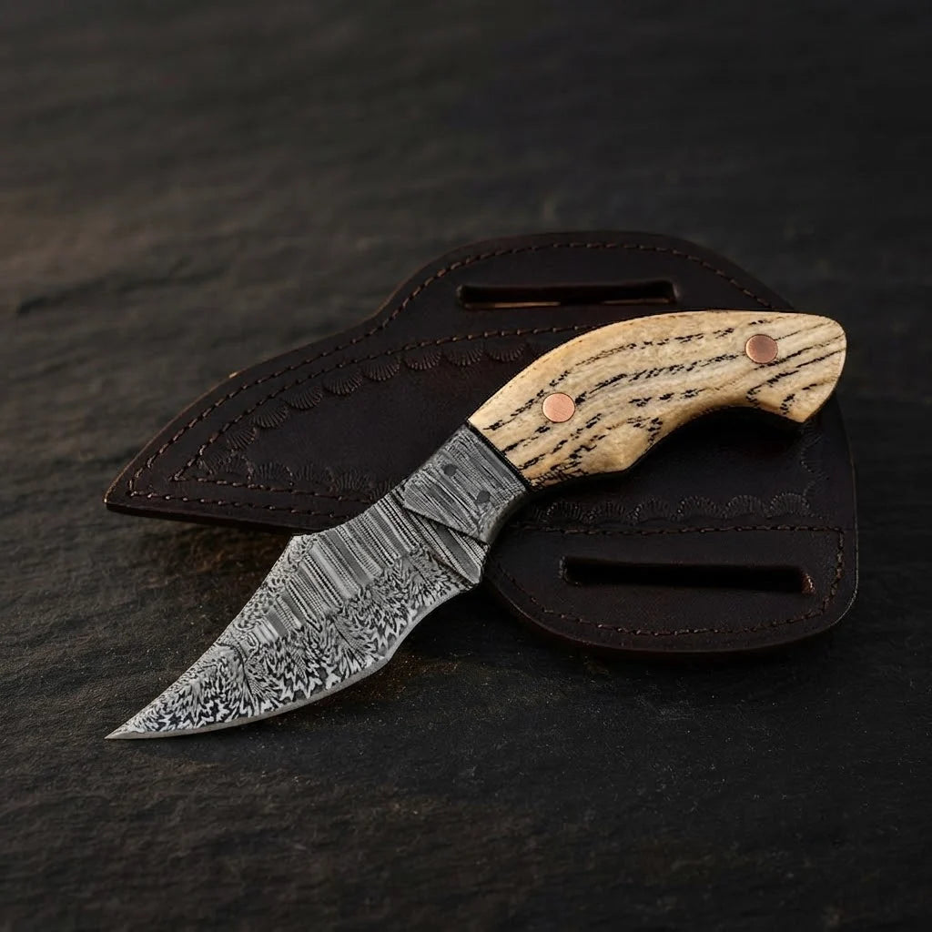 A professional studio shot of a handmade Damascus EDC cowboy knife featuring an ashwood handle with copper pins, Damascus bolster, and leather pancake sheath on a dark stone background