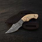 A professional studio shot of a handmade Damascus EDC cowboy knife featuring an ashwood handle with copper pins, Damascus bolster, and leather pancake sheath on a dark stone background