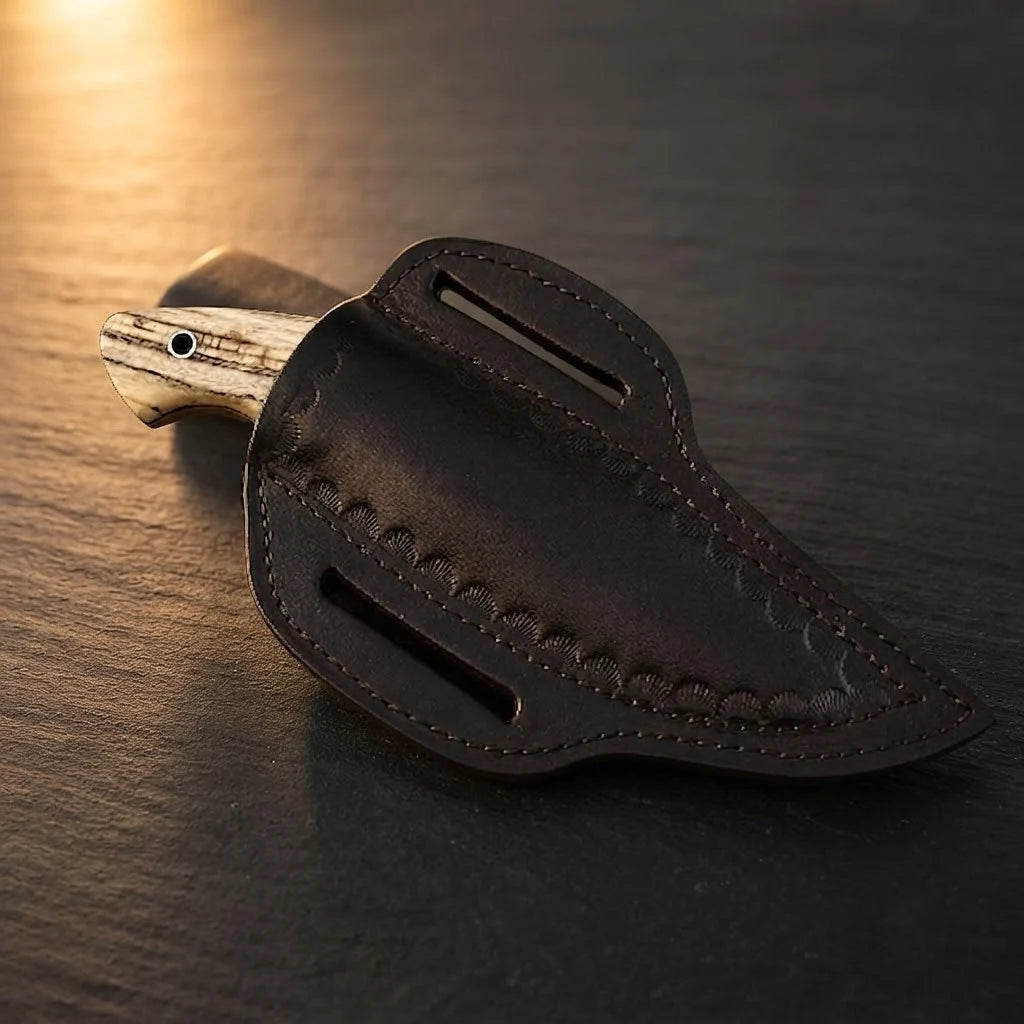 A professional handmade Damascus EDC cowboy knife fully enclosed in its hand-tooled leather pancake sheath, showcasing the ashwood handle pommel on a dark stone background