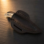A professional handmade Damascus EDC cowboy knife fully enclosed in its hand-tooled leather pancake sheath, showcasing the ashwood handle pommel on a dark stone background