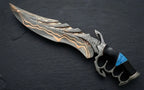 A professional studio shot of a handmade Damascus steel fantasy medieval dagger with a dark skeleton knuckle-duster handle and vibrant turquoise stone inlay resting on a dark slate surface