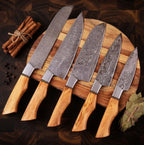 A professional set of hand-forged Damascus steel kitchen knives featuring natural olive wood handles, arranged on a round wooden cutting board with cinnamon sticks and bay leaves.