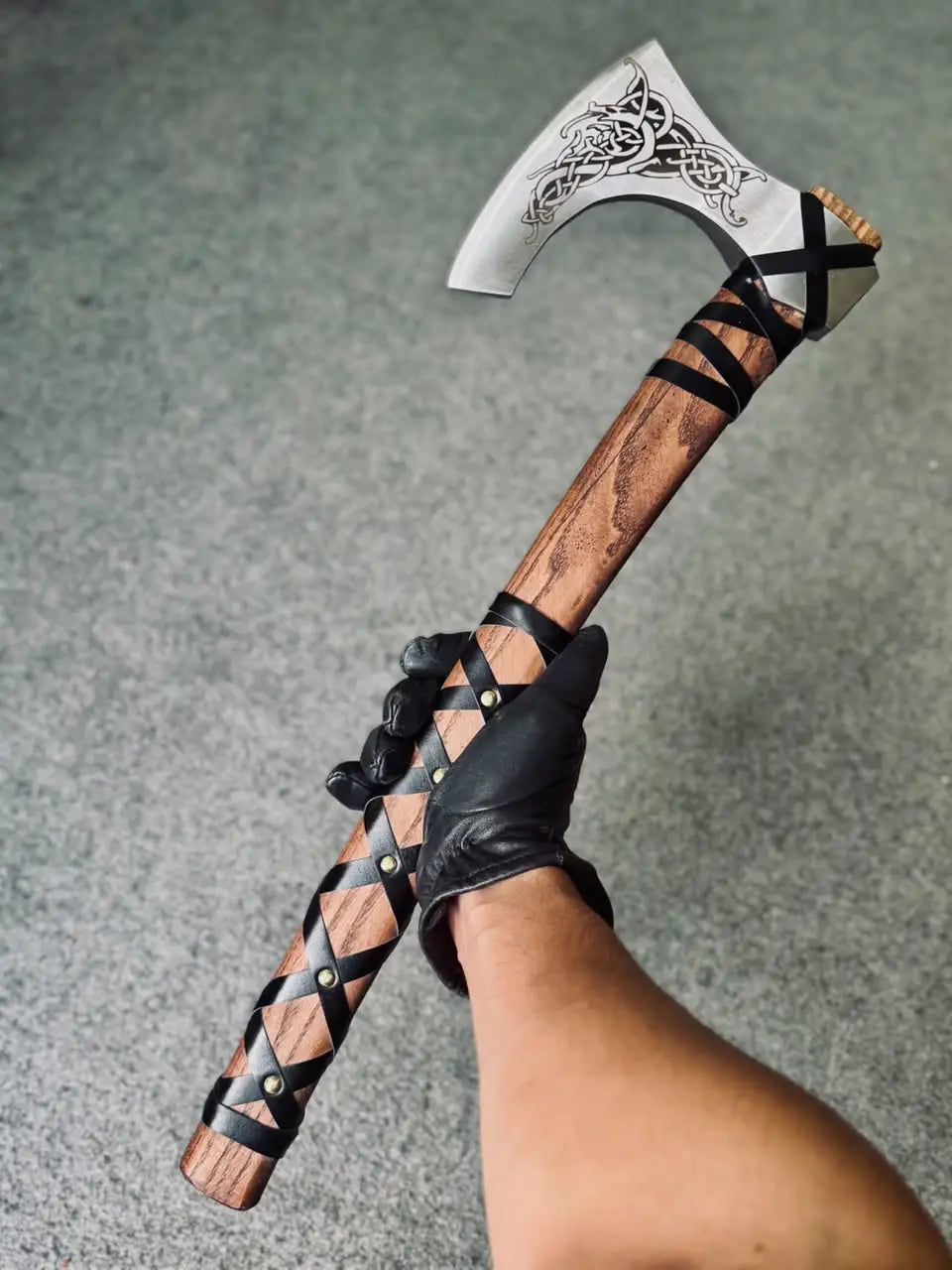 A person wearing a black leather glove holding a handcrafted Viking bearded axe, showing the full length of the wooden handle with its criss-cross leather wrap, brass pins, and the polished steel blade featuring detailed Nordic knotwork engraving.