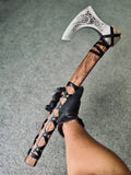 A person wearing a black leather glove holding a handcrafted Viking bearded axe, showing the full length of the wooden handle with its criss-cross leather wrap, brass pins, and the polished steel blade featuring detailed Nordic knotwork engraving.