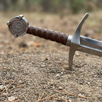 Premium handcrafted Knight Templar sword hilt detail featuring a ribbed leather grip and rustic steel crossguard, captured in a dramatic outdoor setting with a soft-focus natural background.