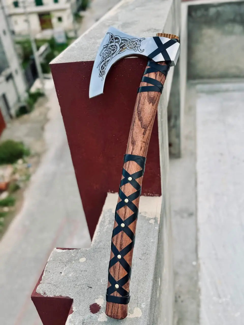 Premium hand-forged custom Viking axe featuring an engraved Norse dragon pattern on a polished bearded blade and an ergonomic leather-wrapped wooden handle, photographed outdoors in daylight.