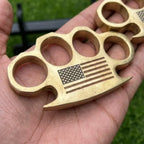 Close-up of an artisan-crafted solid brass paperweight featuring a laser-engraved American flag, held in hand against a natural green backdrop.