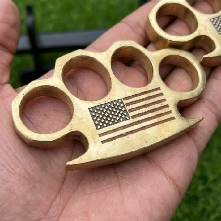 Close-up of an artisan-crafted solid brass paperweight featuring a laser-engraved American flag, held in hand against a natural green backdrop.