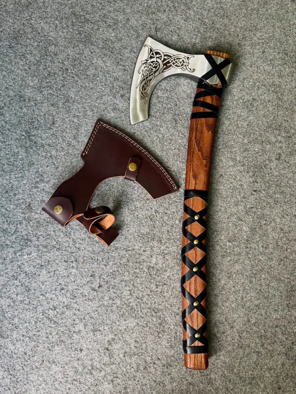 Top-down flat lay view of a handcrafted Viking bearded axe and its matching brown leather sheath on a textured grey background, showcasing the polished steel blade with intricate Nordic knotwork engraving and a criss-cross leather-wrapped wooden handle.