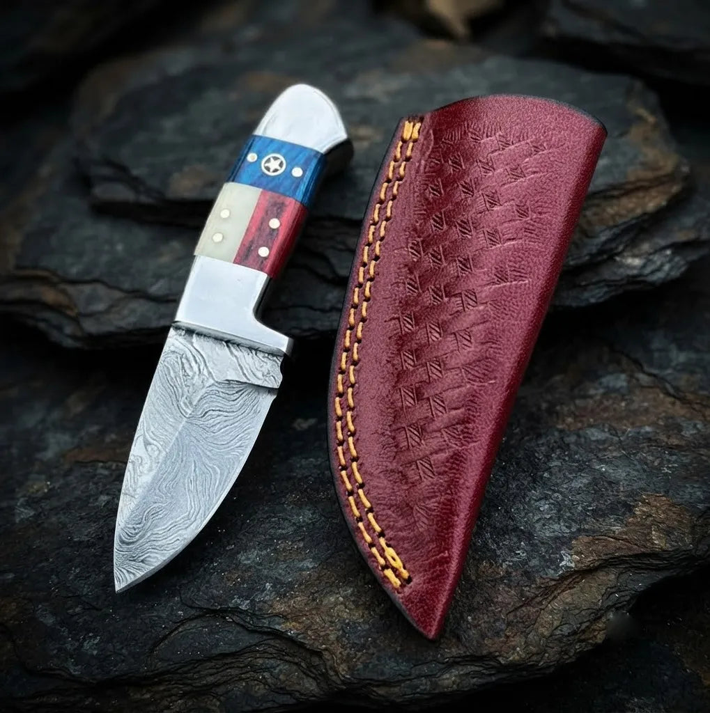 Handmade Damascus steel skinner knife featuring a unique American flag design handle with a star detail, paired with a hand-stitched maroon leather sheath on a dark stone background