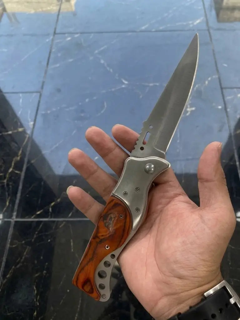 A close-up lifestyle shot of a person's hand holding an open stainless steel folding pocket knife, showcasing the full length of the satin-finish spear point blade with spine jimping, a metallic bolster, and an ergonomic handle scales with polished orange burl wood inlay against a black marble background.