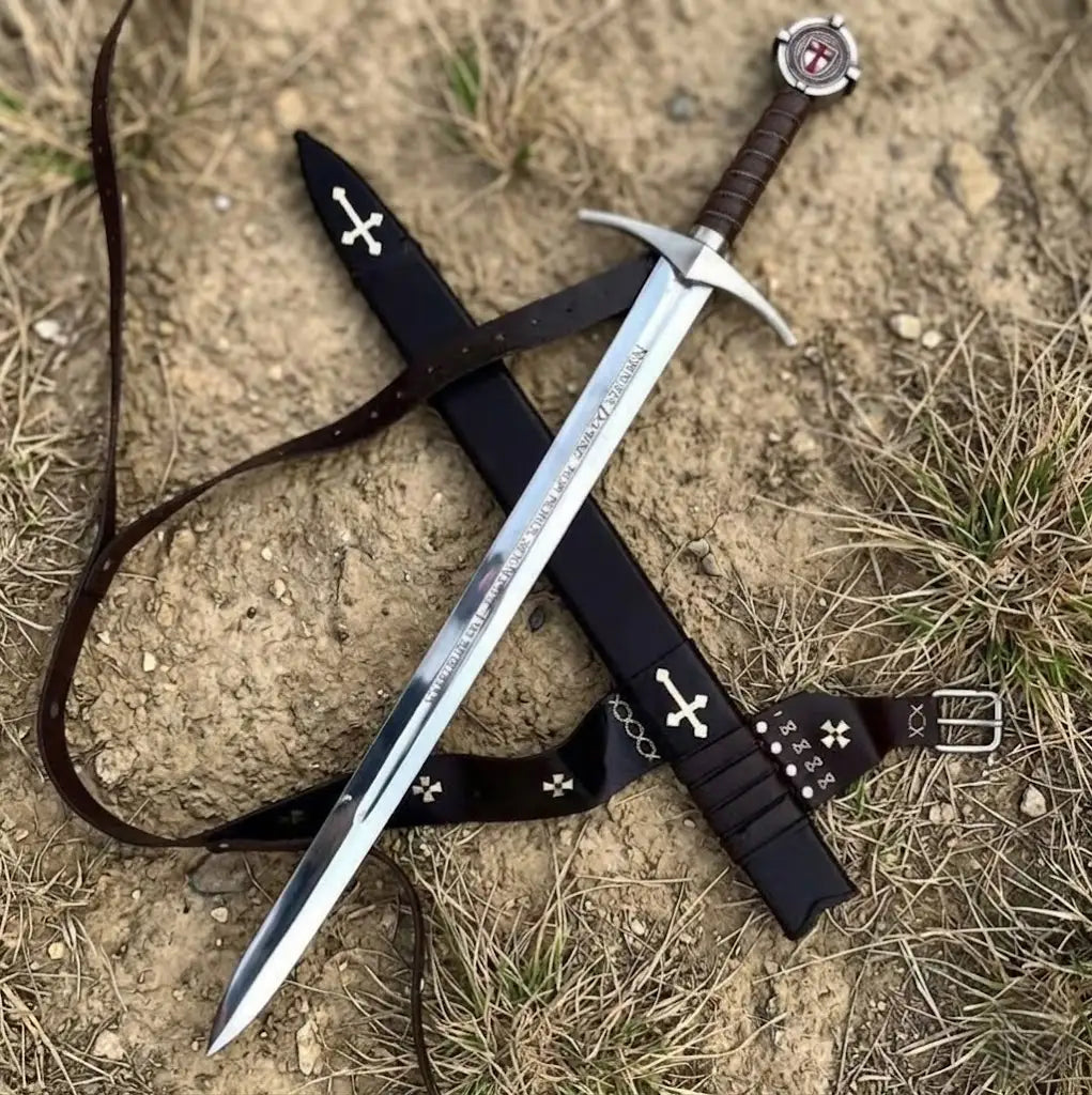 High-angle shot of a handcrafted medieval Knight Templar longsword crossed over its black leather scabbard on dirt ground, featuring a polished steel blade with engraved Latin inscriptions, a leather-wrapped grip, and a red cross emblem on the pommel.