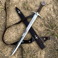 High-angle shot of a handcrafted medieval Knight Templar longsword crossed over its black leather scabbard on dirt ground, featuring a polished steel blade with engraved Latin inscriptions, a leather-wrapped grip, and a red cross emblem on the pommel.
