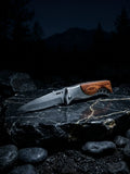 A cinematic outdoor studio shot of a premium stainless steel folding pocket knife resting open on a polished black marble stone with gold veins at night, showcasing a brushed steel blade with jimping, a metallic bolster, and a rich curved wood grain inlay handle.