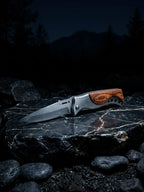 A cinematic outdoor studio shot of a premium stainless steel folding pocket knife resting open on a polished black marble stone with gold veins at night, showcasing a brushed steel blade with jimping, a metallic bolster, and a rich curved wood grain inlay handle.