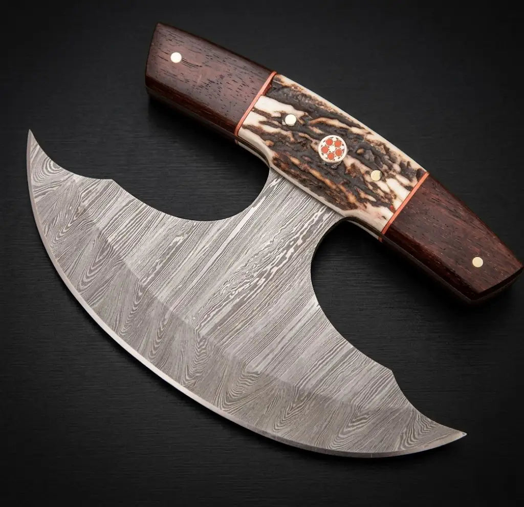 A top-down flat lay view of a premium handcrafted Damascus steel ulu push dagger knife on a clean black background, showcasing the beautiful wavy twist pattern of the layered steel blade, a highly textured stag antler center handle with an intricate red mosaic pin, dark wooden bolsters, and copper liners.