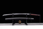 Handcrafted black Damascus steel katana sword displayed on a black wooden stand, featuring a blacked-out blade, red and black wrapped hilt, and matching textured scabbard.