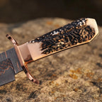 Close-up of a handmade Copper Damascus Bowie knife, showcasing the textured natural stag antler handle, polished copper guard, and copper-layered steel on a stone background
