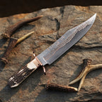 A professional handmade Copper Damascus Bowie knife displayed on a rugged stone surface, showcasing its natural antler handle, copper guard, and intricate blade pattern
