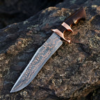 Close-up of a handmade Copper Damascus Bowie knife, showcasing the unique copper-layered steel pattern, double copper guard, and rich wood handle on a stone background
