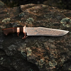 A professional handmade Copper Damascus Bowie knife displayed horizontally, showcasing its full blade profile, double copper guard, and wood handle on a textured stone surface