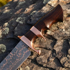 Close-up of a handmade Copper Damascus Bowie knife, showcasing the intricate double copper guard, ergonomic wood handle, and copper-layered blade on a textured stone background