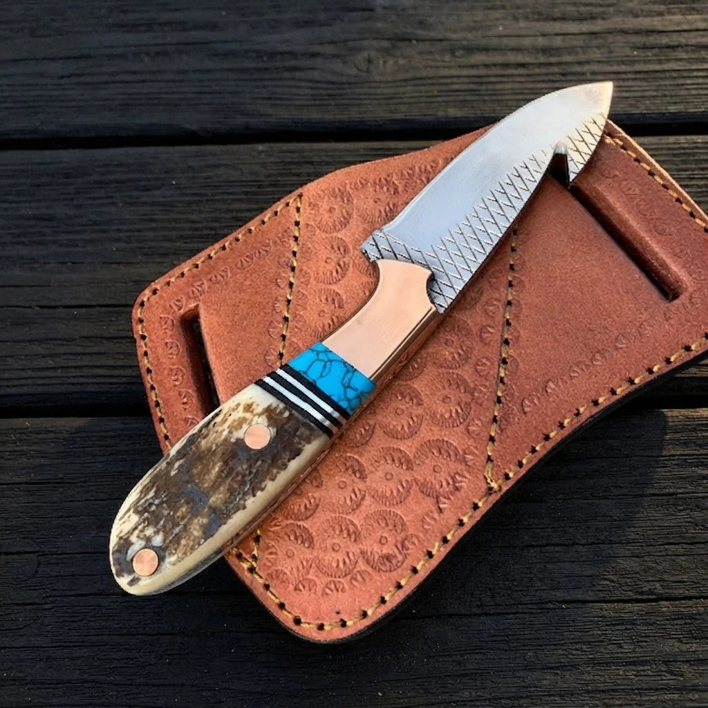 A professional angled studio view of a handmade D2 steel cowboy gut hook knife with a copper bolster, stag antler handle, and turquoise spacers resting on a pancake leather sheath