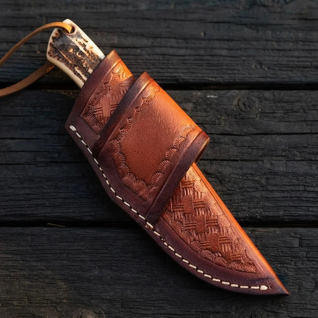 A professional handmade D2 steel cowboy knife securely sheathed in a hand-tooled leather holster, showcasing the stag antler handle pommel and lanyard on a dark wooden background.