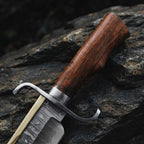 Close-up of a handmade Damascus Bowie knife, showcasing the rich-grained Rosewood handle, solid metal bolster, and intricate wavy Damascus steel pattern on the blade