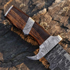 Close-up of a handmade Damascus Bowie knife, showcasing the rich-grained Wenge wood handle, intricate Damascus steel bolster, and finger guard against a dark stone background.