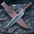 A professional handmade Damascus Bowie knife featuring a VG10 steel blade and Rosewood handle, paired with its custom-fitted leather sheath on a textured stone surface.