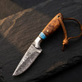 A professional handmade Damascus cowboy hunting knife featuring a rosewood handle, vibrant turquoise spacer, and a leather lanyard, displayed on a textured stone surface