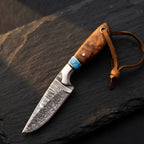 A professional handmade Damascus cowboy hunting knife featuring a rosewood handle, vibrant turquoise spacer, and a leather lanyard, displayed on a textured stone surface