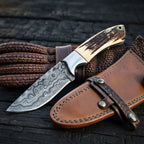 A professional handmade Damascus steel cowboy knife featuring a raindrop pattern blade and stag antler handle, displayed with its button-lock leather sheath on a dark textured background