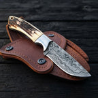 A professional handmade Damascus steel cowboy knife resting on its custom leather snap-button holster, showcasing the intricate steel pattern and stag antler handle on a dark wooden background