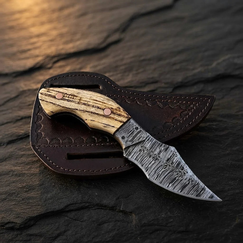 A professional handmade Damascus EDC cowboy knife resting on its leather pancake sheath, showcasing the ashwood handle, copper pins, and patterned blade on a dark stone surface